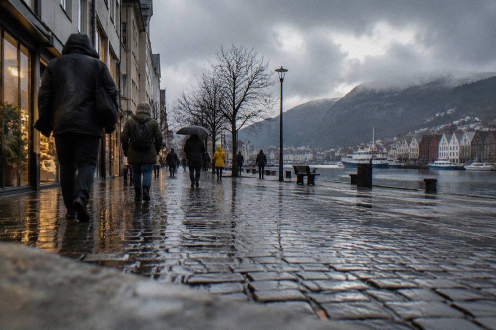 Rain-slicked cobblestone street in central Bergen with people in rain jackets walking past lit shop windows