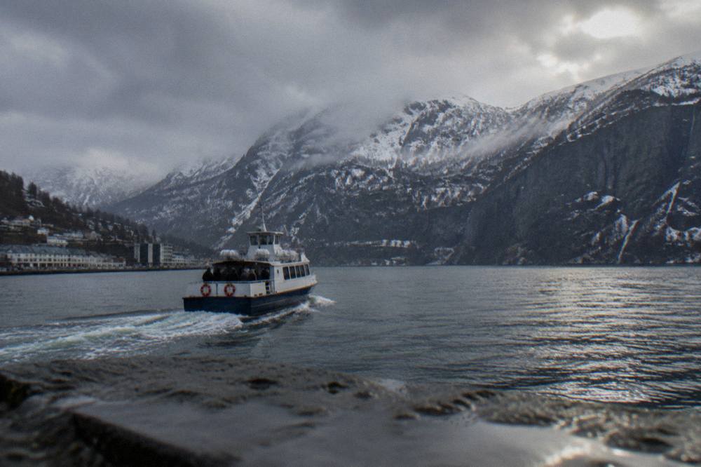 A small passenger boat cutting across dark fjord water toward steep snow-dusted cliffs in winter