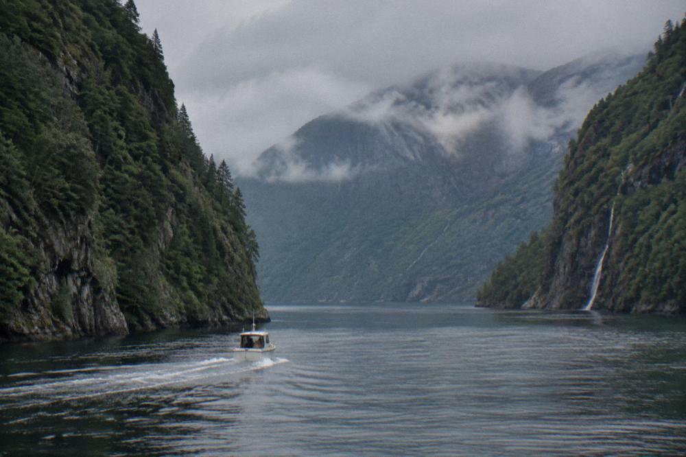 Small boat cruising a narrow fjord between steep forested cliffs in western Norway