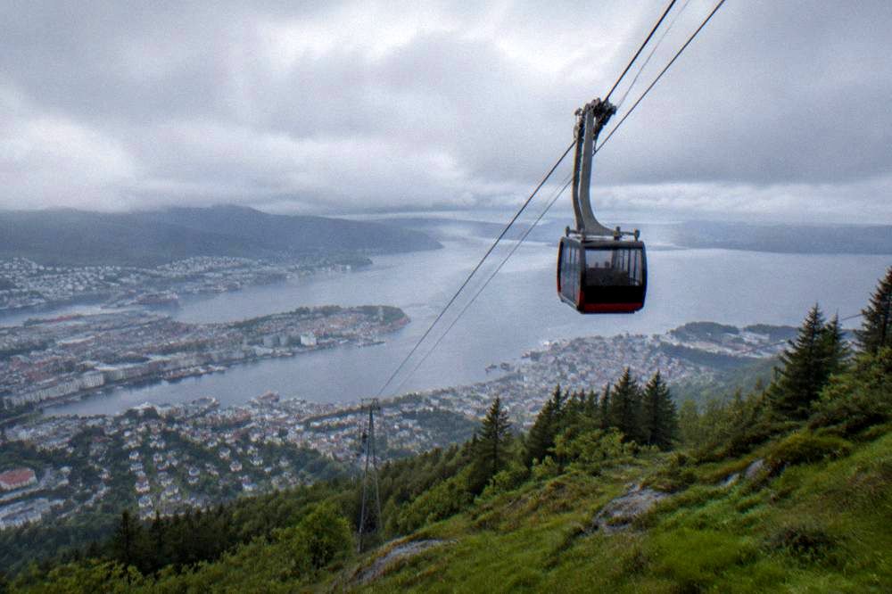 A cable car ascending a green mountain above Bergen with the city and fjord visible below
