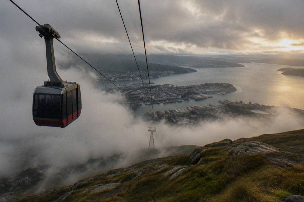 Cable car ascending above clouds toward a rocky mountain plateau overlooking fjords