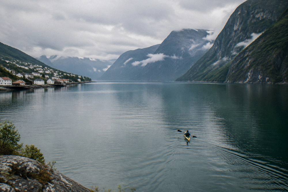 Kayaker paddling on glassy fjord water beneath steep mountain walls at dusk