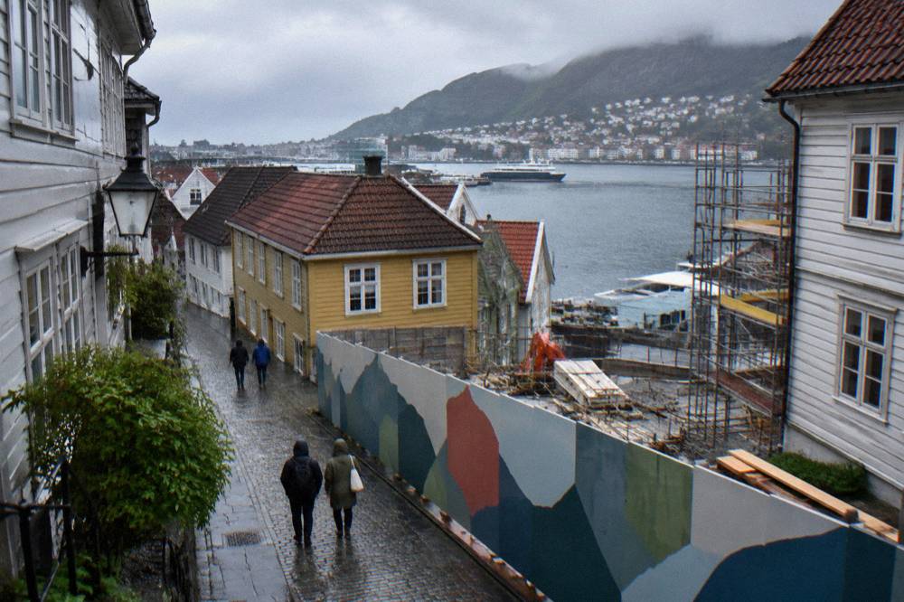 A pedestrian lane in central Bergen with painted hoarding fencing off a construction site beside old timber buildings.
