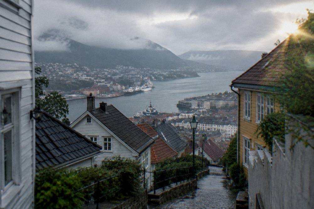 A view over Bergen's timber rooftops toward the harbour from a hillside path, with painted walls in the foreground streets.