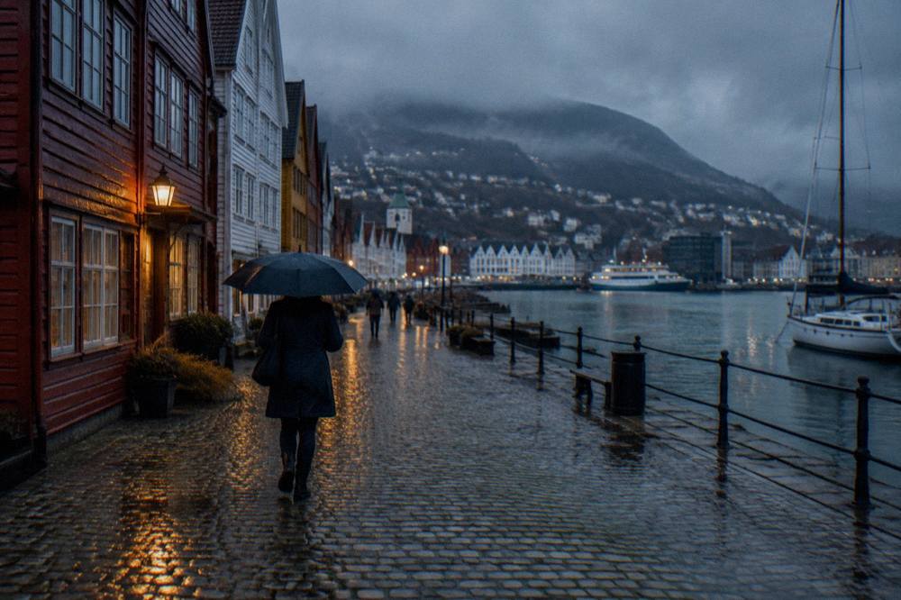 A person with an umbrella walking across cobblestones in old Bergen while light rain falls