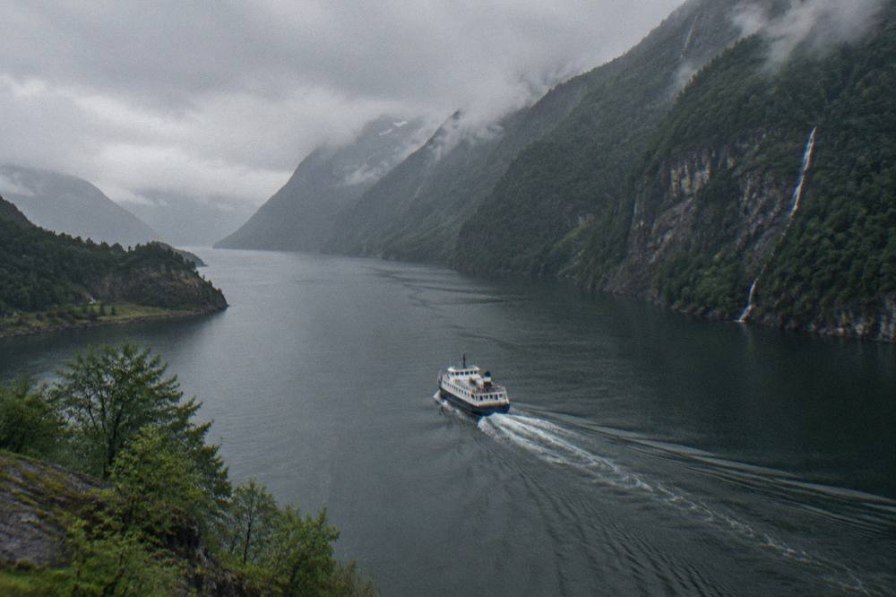 A fjord boat cutting through light rain across dark water with steep forested cliffs on both sides