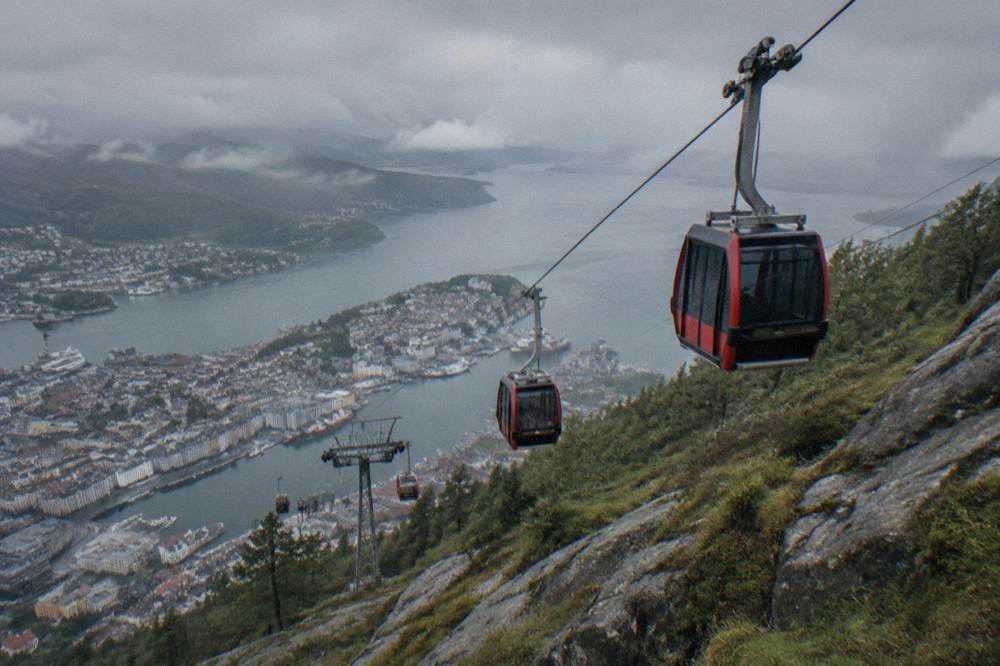 Cable car cabins travelling up a steep rocky mountainside above the city of Bergen with the fjord in the background