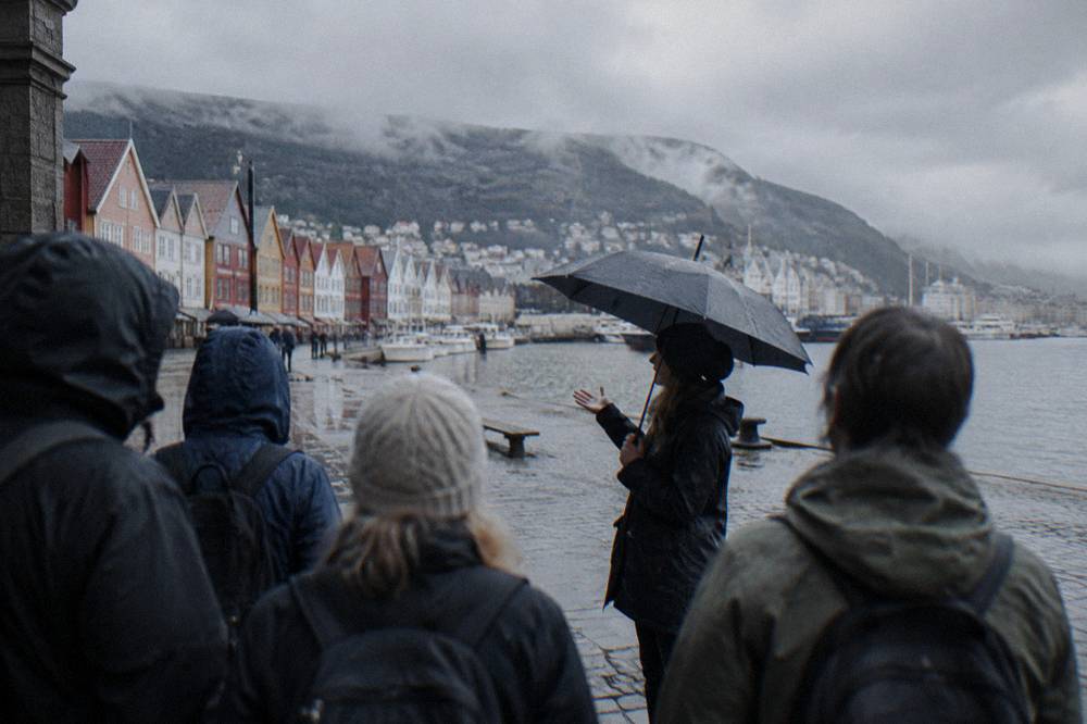 A tour guide holding an umbrella while speaking to a small group of visitors on a wet cobblestone street in Bergen