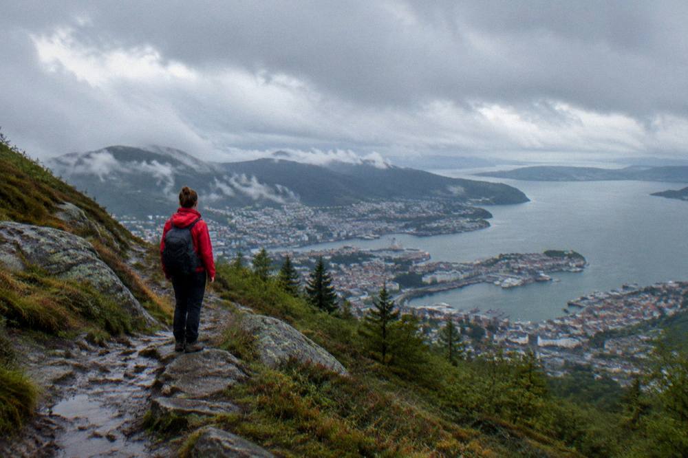 Hiker in a red jacket standing on a mountain trail above Bergen with the city and fjord visible far below