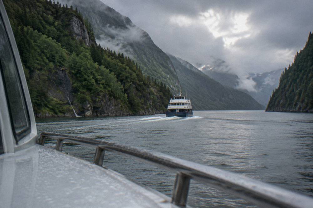 A small passenger boat crossing a narrow fjord channel with steep forested cliffs on both sides