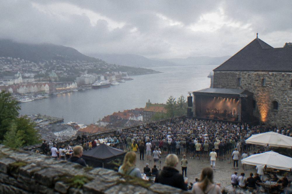 A festival crowd gathered outside a stone fortress venue beside the harbour in Bergen on a summer evening