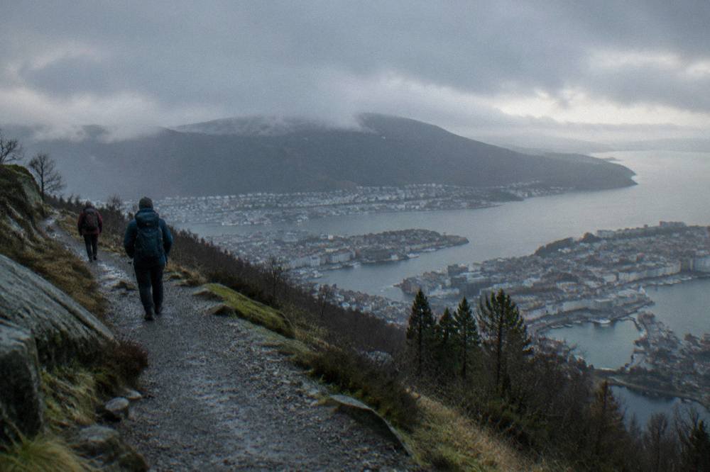 Two hikers with backpacks on a gravel trail above the city of Bergen with the harbour far below