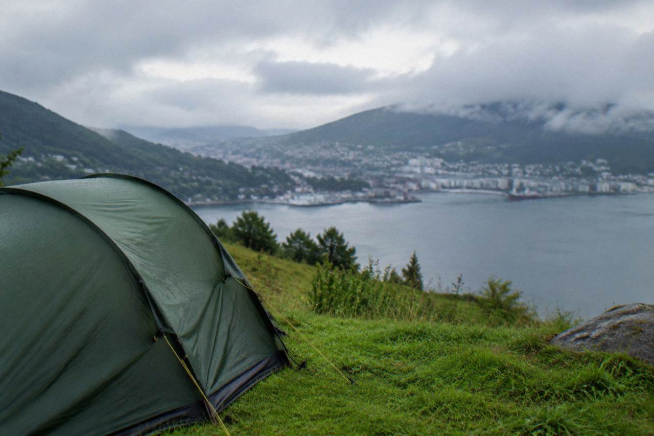 A tent pitched on green grass with a view over a fjord and forested hills