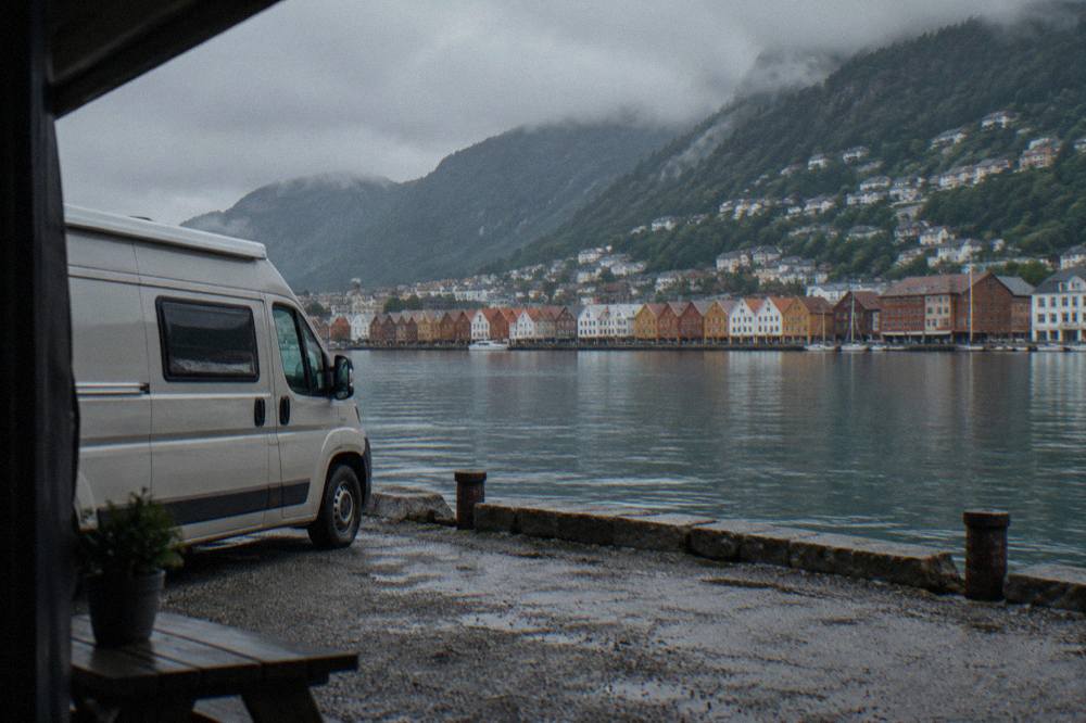 A campervan parked beside a calm fjord with wooden houses on the far shore