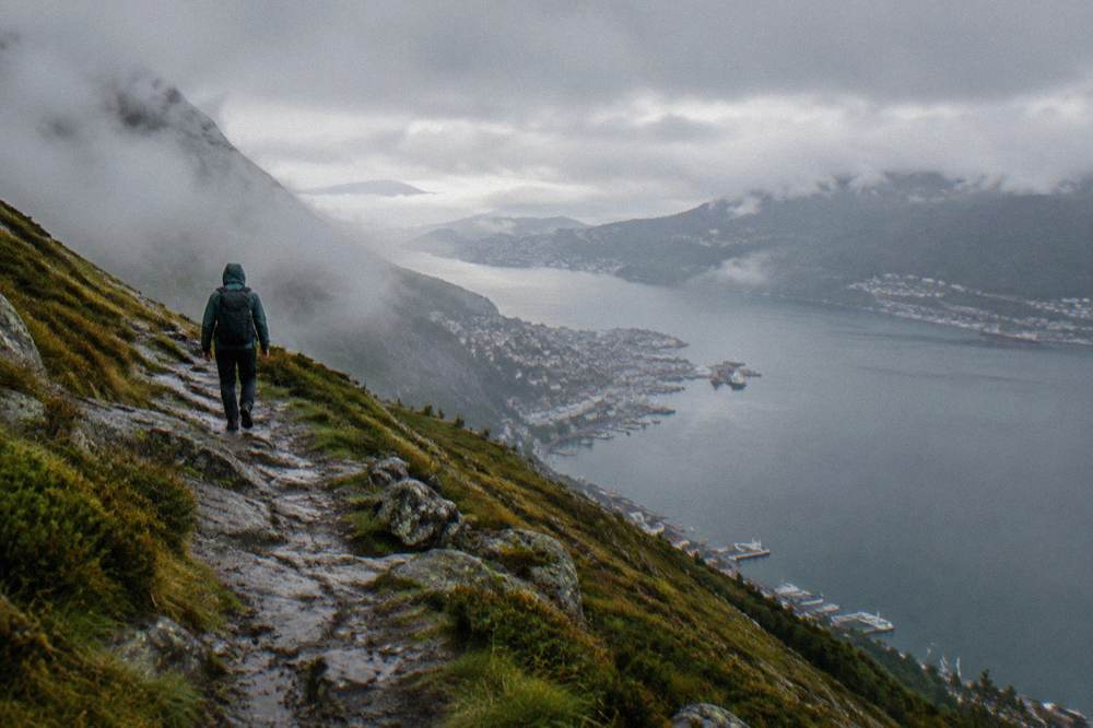 A hiker with a backpack walking a rocky trail above a fjord in low cloud