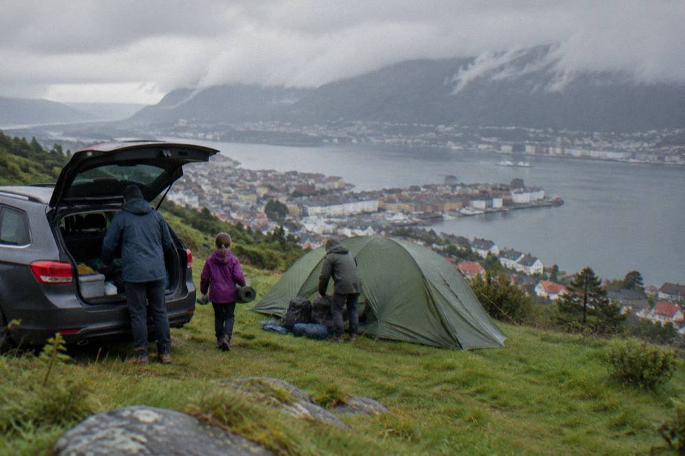A family unloading camping gear from a car beside a tent in a green field