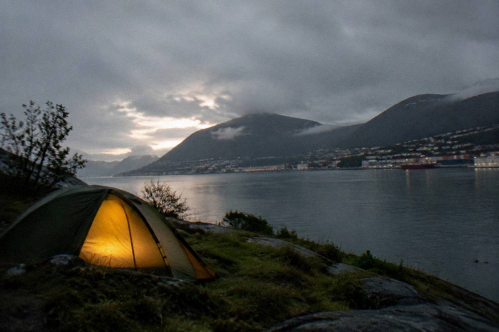 A tent glowing from inside at sunset with mountains silhouetted across a fjord