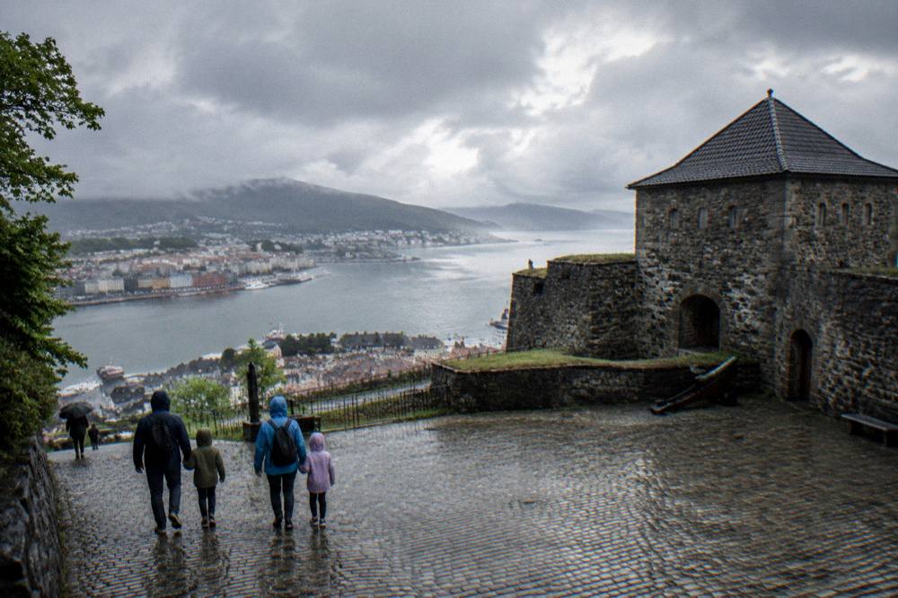 A family with rain jackets walking across a cobblestone square near an old stone fortress