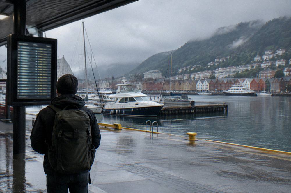 A traveller with a backpack studying a departures board beside moored boats at a harbour terminal