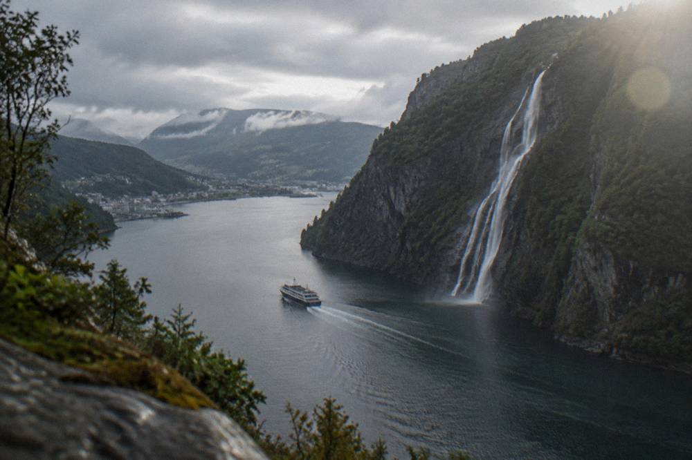 A small cruise boat passing through a narrow strait with water pouring down a cliff face