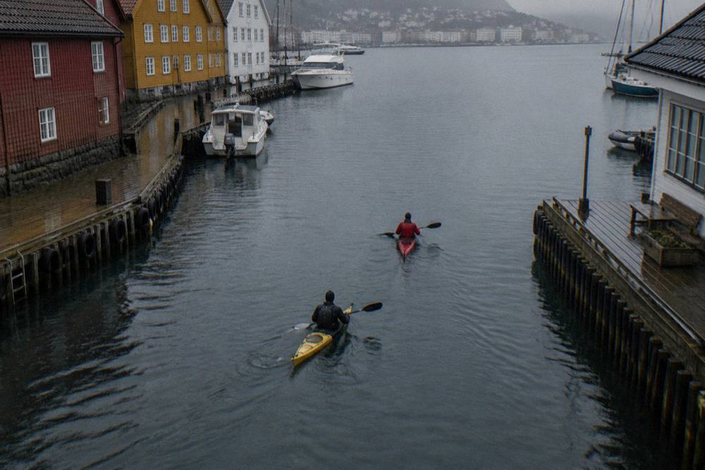 Two kayakers paddling on calm water near wooden docks in a Norwegian harbour