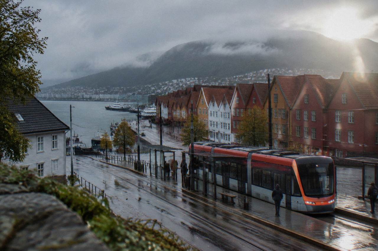 A modern light rail tram waiting at a street stop in central Bergen with colourful wooden buildings behind it