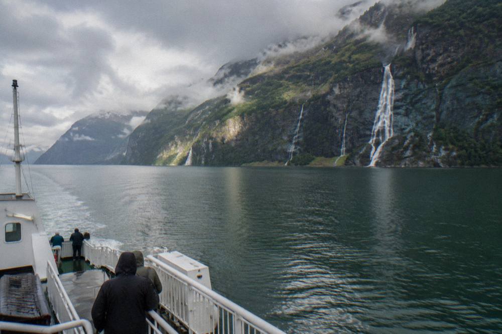 A ferry crossing green fjord water toward steep waterfall cliffs in the Hardangerfjord region