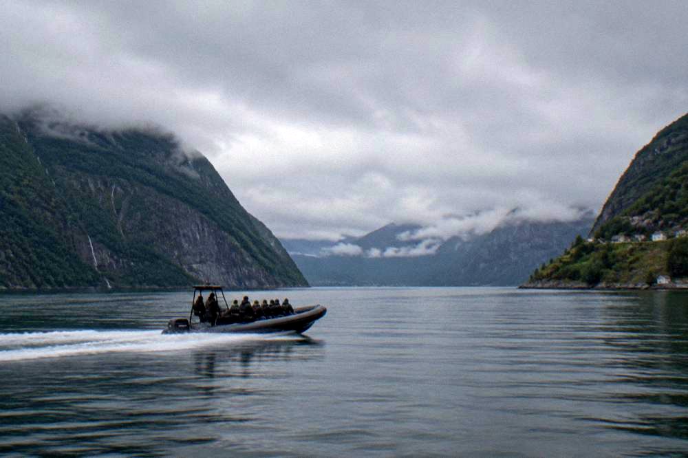 A rigid inflatable speedboat cutting across calm fjord water between steep green cliffs