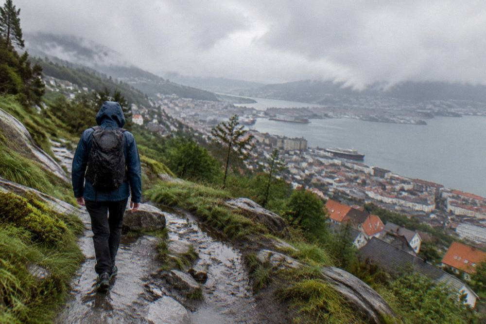 A hiker in waterproof jacket walking a wet mountain trail above Bergen's rooftops