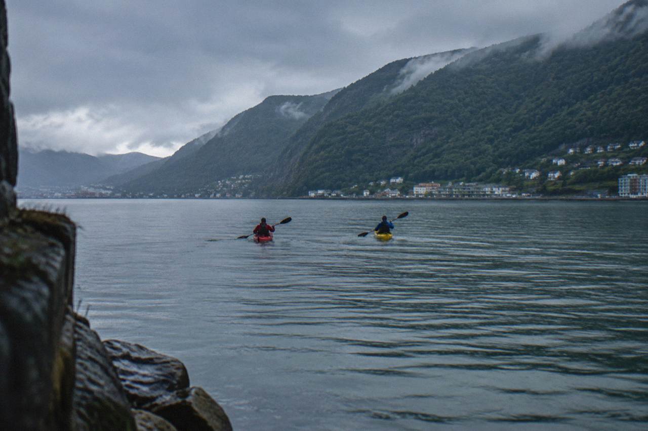 Two kayakers paddling across calm grey-green fjord water with steep forested mountains rising behind them near Bergen