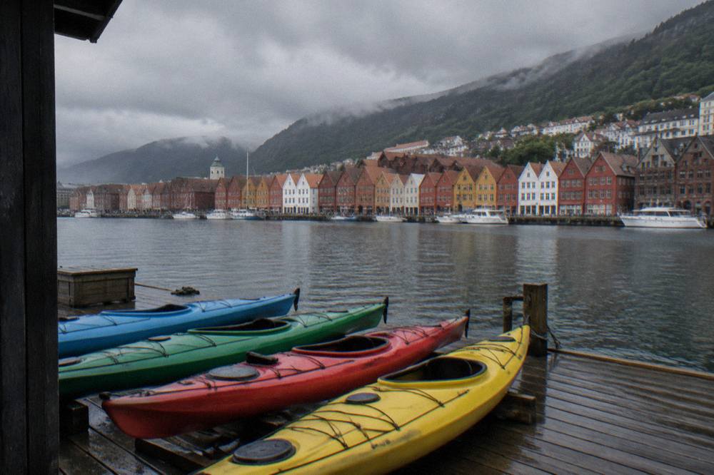 A row of kayaks resting on a wooden jetty with colourful historic harbour buildings across the water