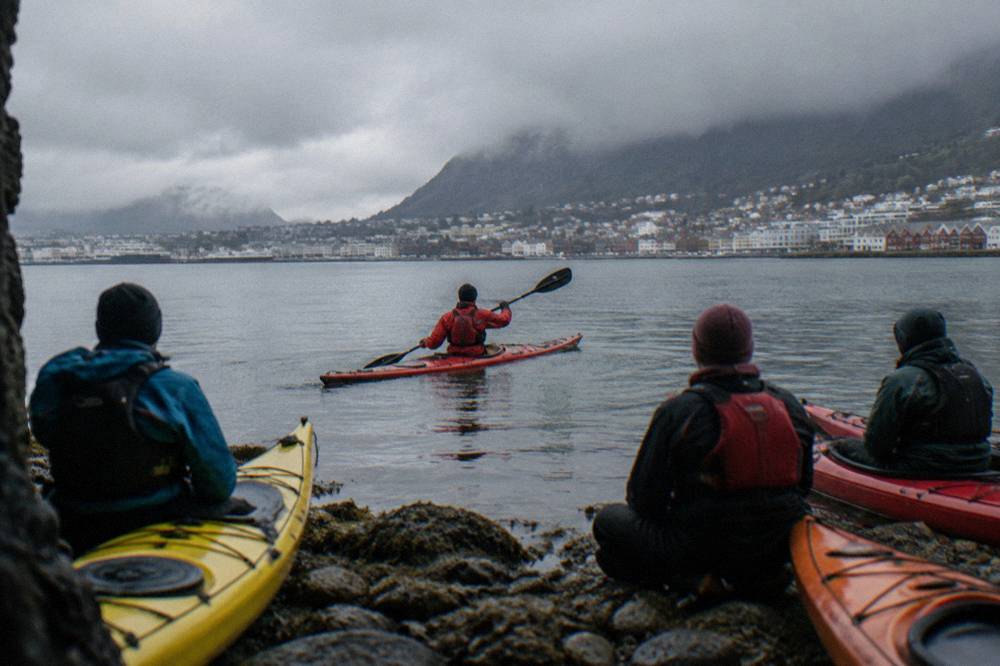 A guide in a red drysuit demonstrating paddle strokes to a small group of kayakers on a rocky shoreline