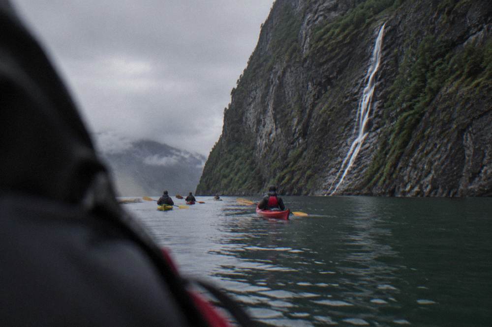 A kayak group passing a steep cliff face streaked with a thin waterfall inside a narrow fjord arm