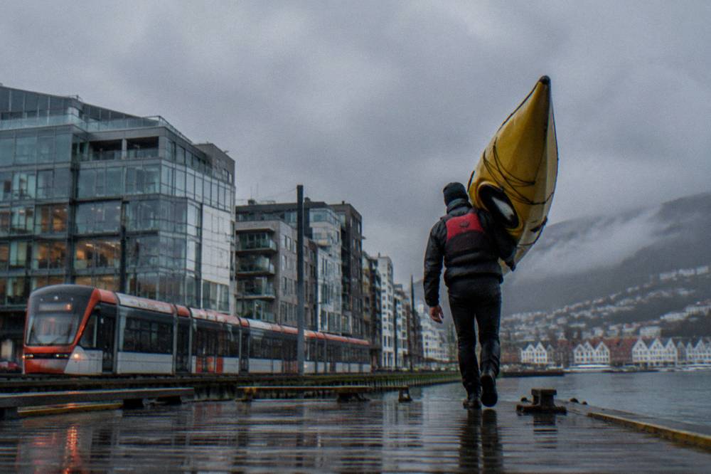 A kayaker carrying a boat on a dock in front of a light-rail tram passing modern waterfront buildings