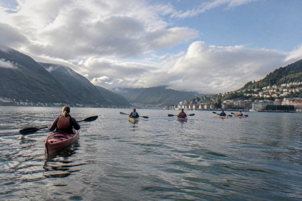 A group of sea kayaks crossing a wide fjord under a bright summer sky with scattered clouds