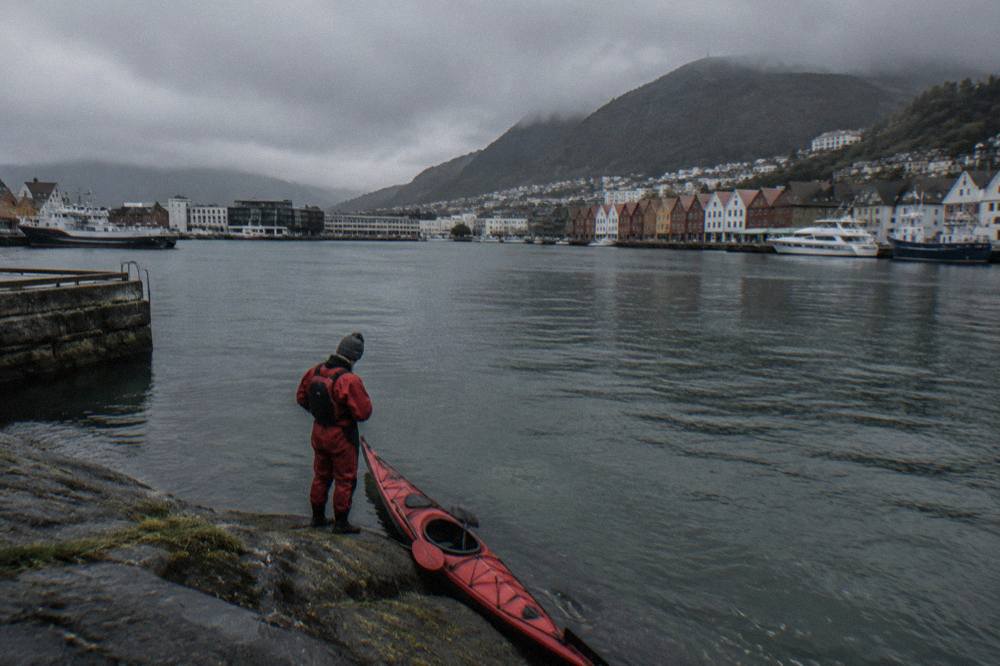 A paddler in a red drysuit and wool hat checking a map beside a kayak loaded with dry bags