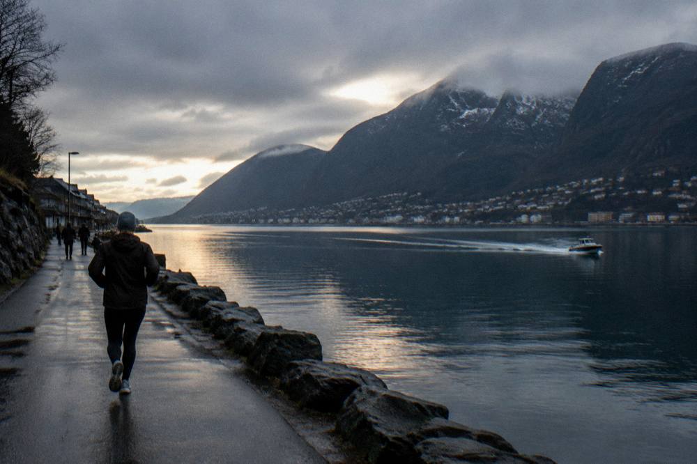 A calm fjord at dusk reflecting steep dark mountains, with a small boat wake crossing the water