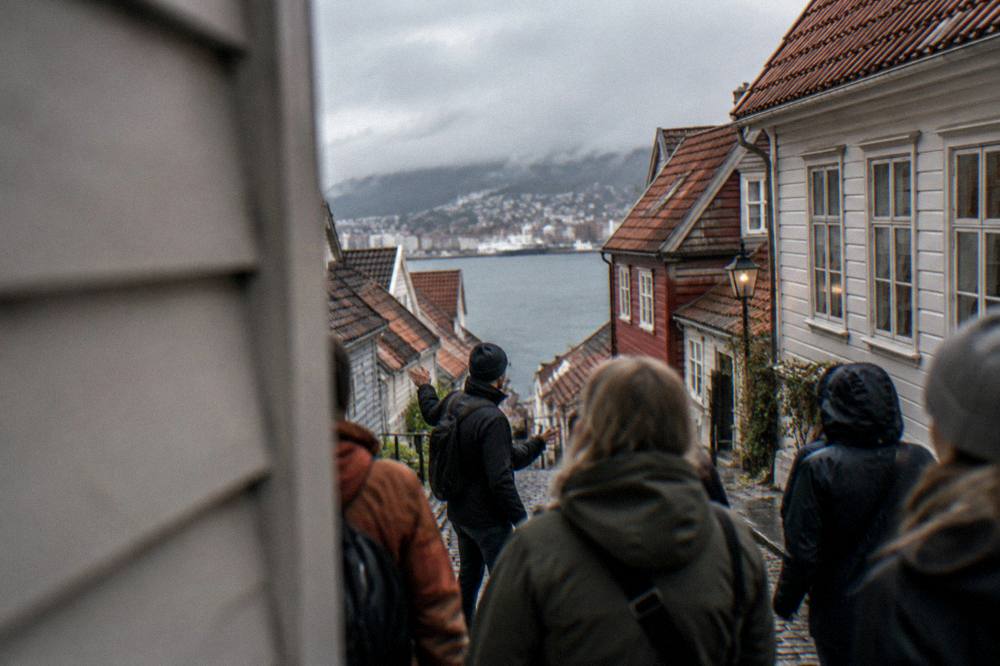 A walking tour group listening to a guide in a narrow Bergen street between wooden houses