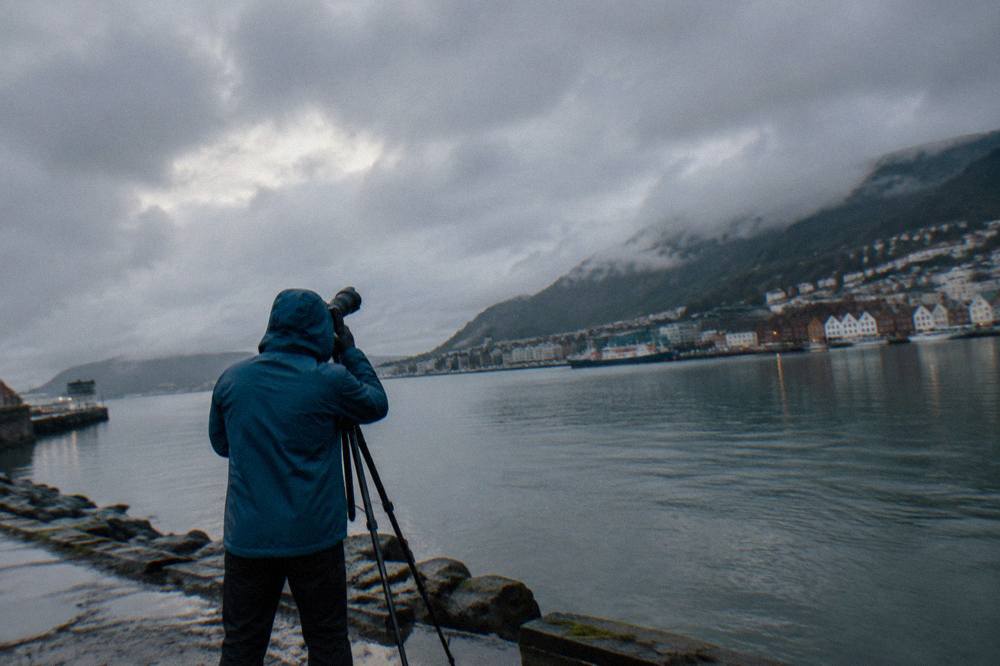 A person in a rain jacket photographing the night sky with a tripod beside a fjord