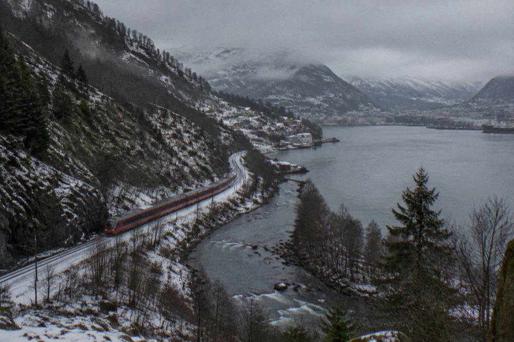 A red train winding along a snow-dusted valley with a river beside the tracks