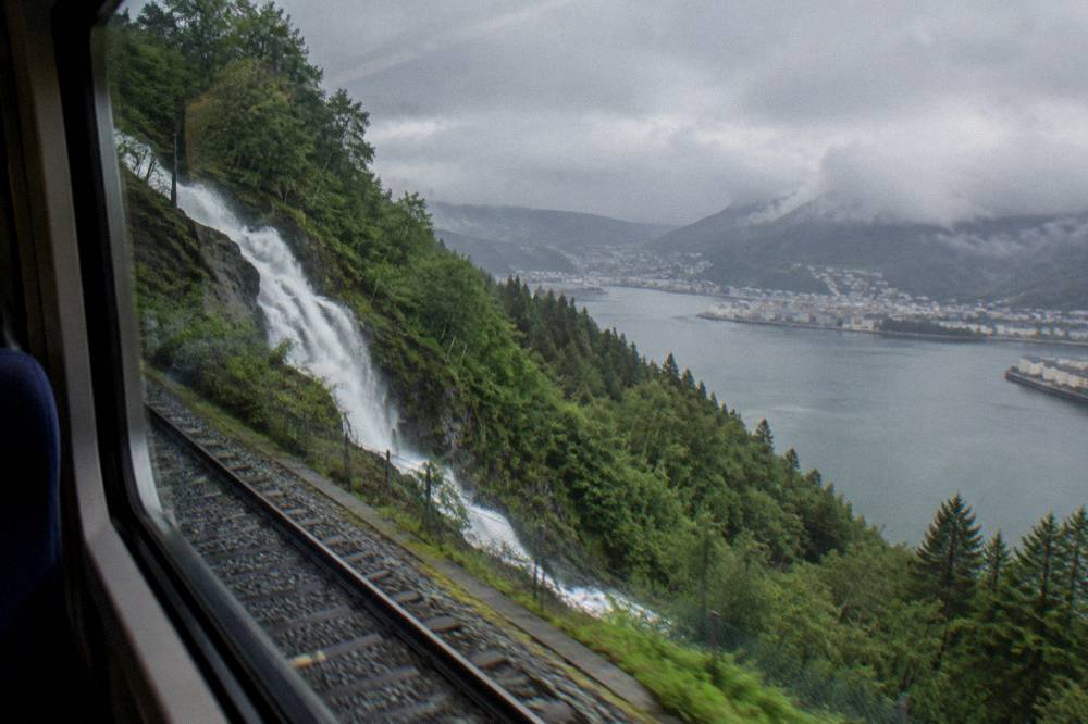 A train window framing a steep green valley with a waterfall falling beside the tracks