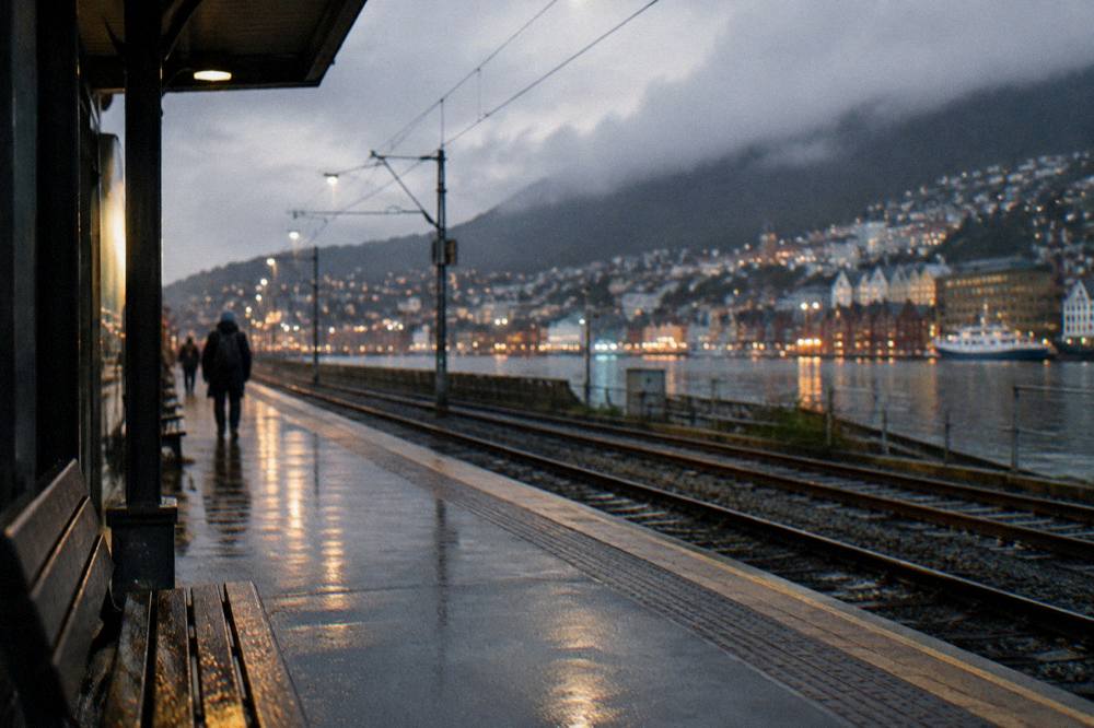 A quiet train platform in Bergen at dusk with the city lights coming on behind the tracks