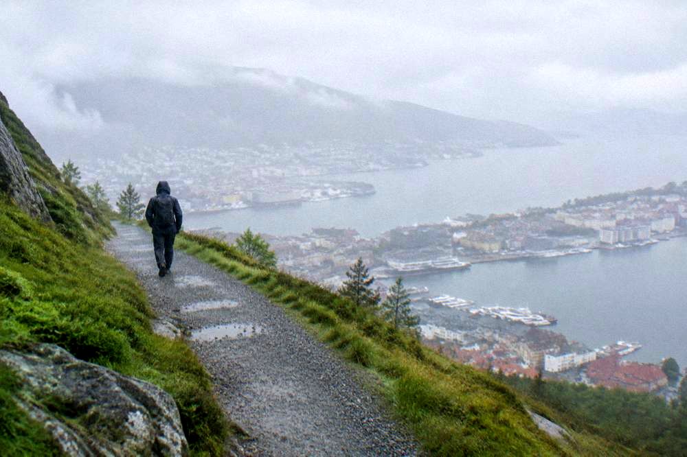 A hiker in rain gear walking a gravel trail above a Norwegian city with water below