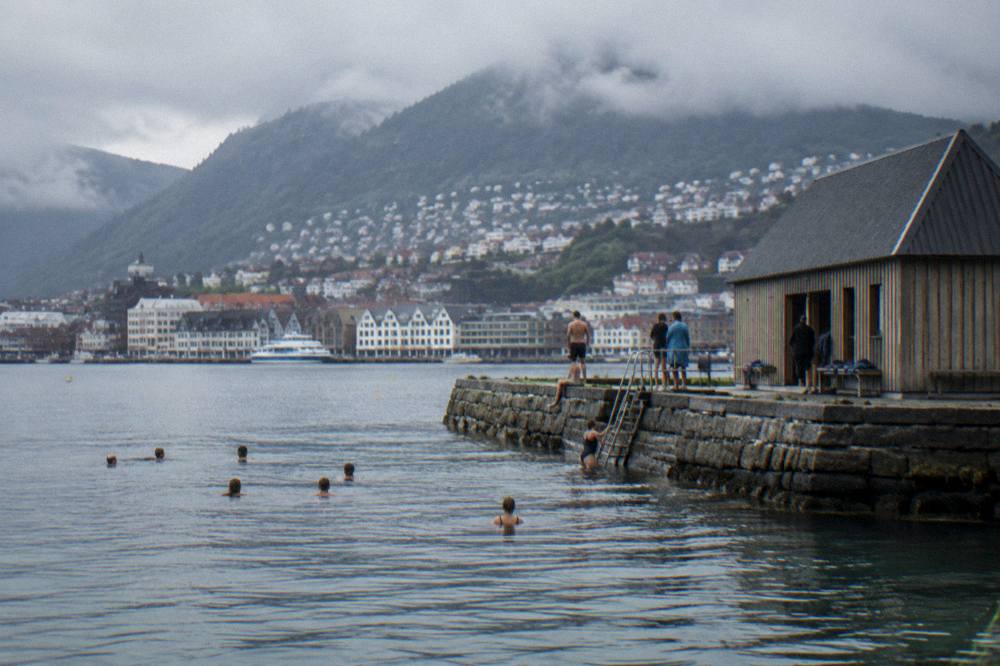 A stone jetty and wooden bathing house at the water's edge with people swimming in cold water