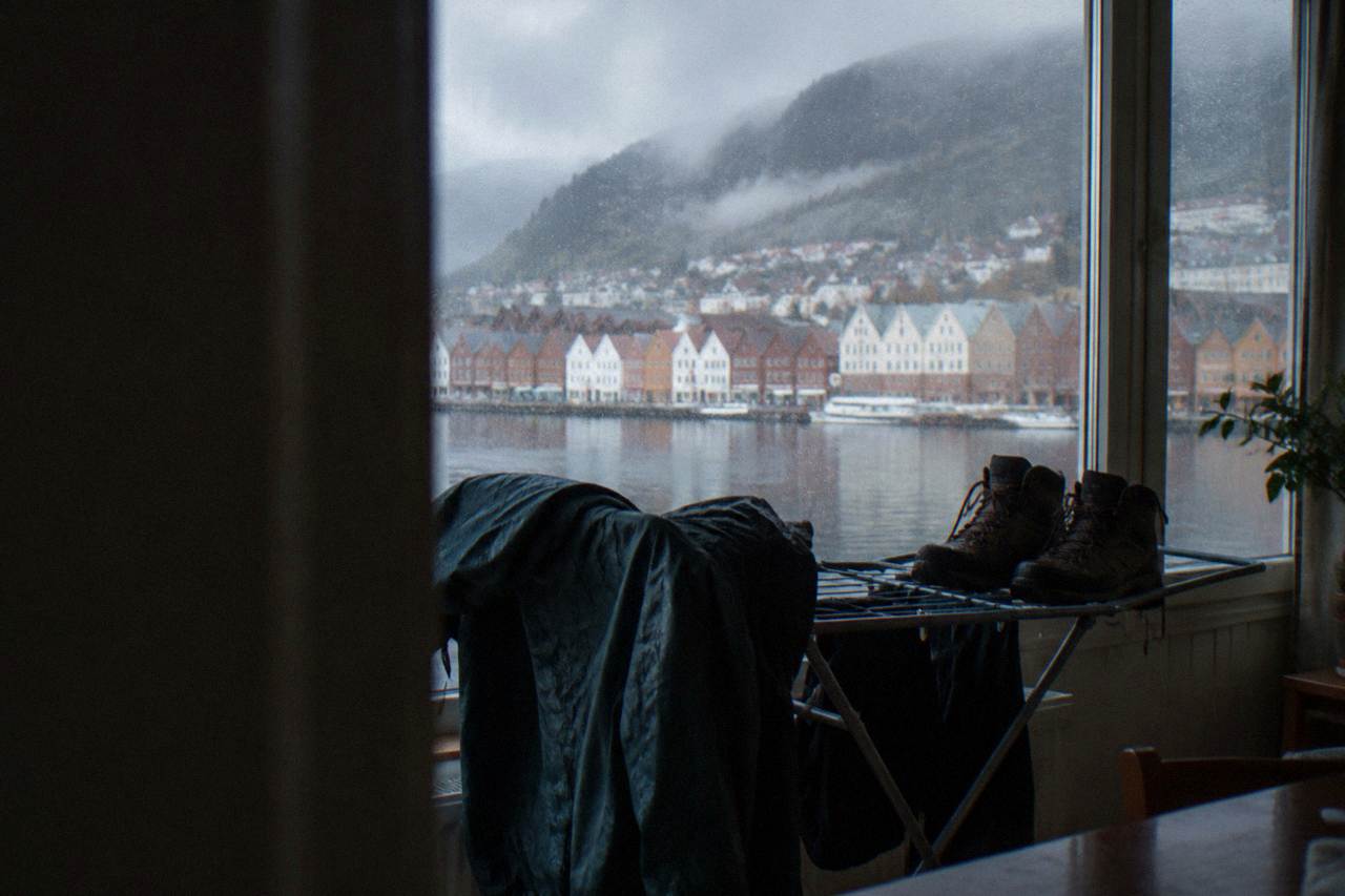 A waterproof jacket and hiking boots drying on a rack beside a window overlooking Bergen's colorful wooden houses in the rain