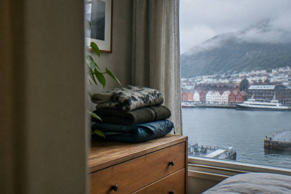 A folded stack of layered clothing including a wool sweater, fleece jacket and waterproof shell on a wooden dresser