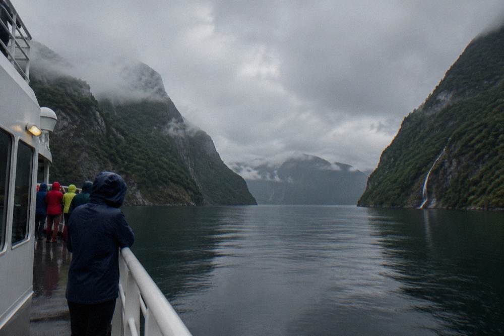A fjord cruise boat on calm dark water with steep forested cliffs rising on both sides and passengers in rain jackets along the rail