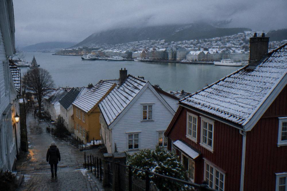 A winter street scene in Bergen with snow dusting the roofs of wooden buildings and a person in an insulated jacket walking past