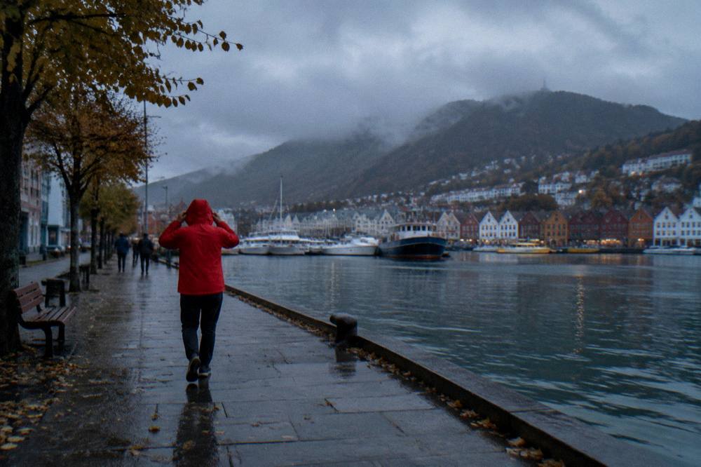 A traveler adjusting the hood of a red rain jacket while looking out over Bergen harbor on an overcast autumn day