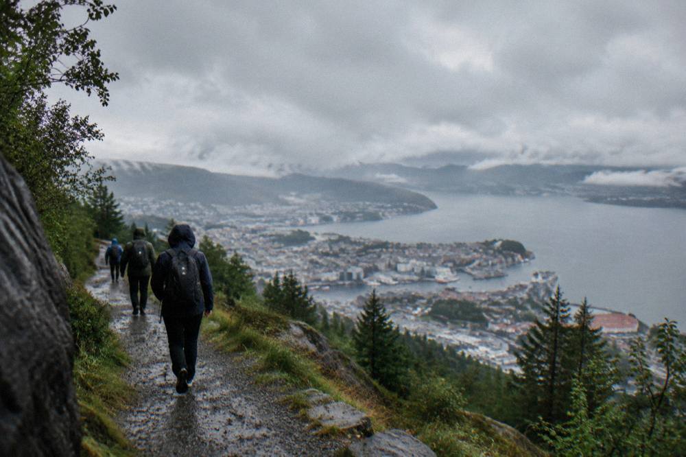 Hikers with backpacks on a gravel trail above Bergen with the city and fjord visible below under grey clouds